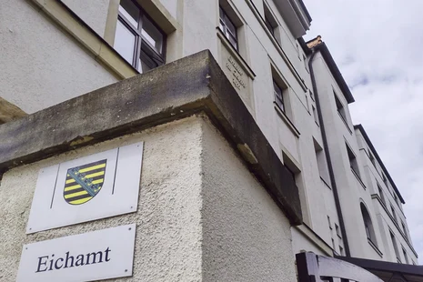 Eingang Eichamt Chemnitz Schild mit Sachsen-Wappen und darunter der Aufschrift „Eichamt“. Im Hintergrund sieht man das helle Gebäude des Eichamtes Chemnitz.
