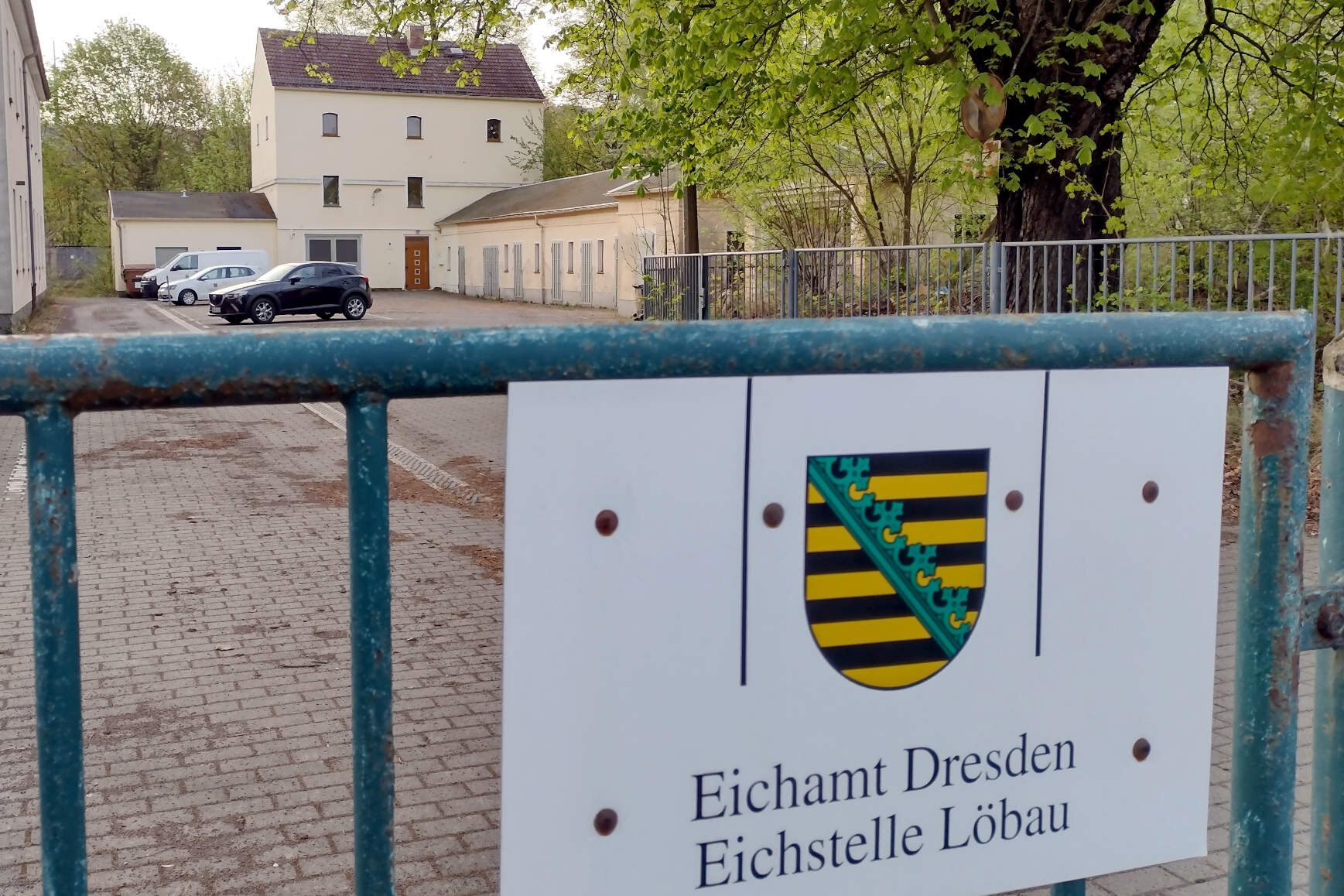 Einfahrt zur Eichstelle Löbau: Am Tor sieht man ein Schild mit Sachsen-Wappen und Aufschrift „Eichamt Dresden, Eichstelle Löbau“. Im Hintergrund sieht man das Gebäude sowie einige Dienstfahrzeuge.