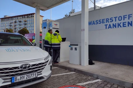 Eichrechtliche Überprüfung einer Wasserstofftankstelle Zwei Personen in Arbeitskleidung an einer Tankstelle, eine tippt auf einem Laptop. An der Zapfsäule steht „H2“. Links ein Prüffahrzeug der Firma H2 Mobility zur Eichung der Wasserstofftankstelle. Im Hintergrund an der Wand: „Wasserstoff tanken“.