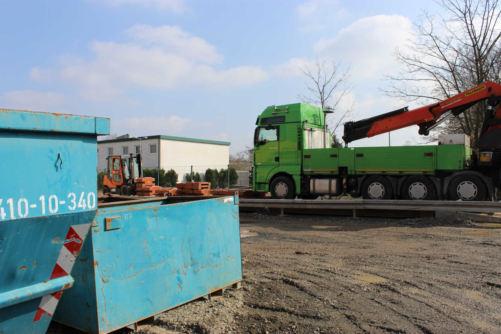 Zwei hellblaue Schrott-Container auf einem Schrottplatz. Der Boden ist erdig und etwas zerfurcht. Im Hintergrund steht ein grüner LKW auf einer Straßenfahrzeugwaage. Ein Gabelstapler stellt große, orangefarbene Prüfgewichte vor den LKW auf die Waage.