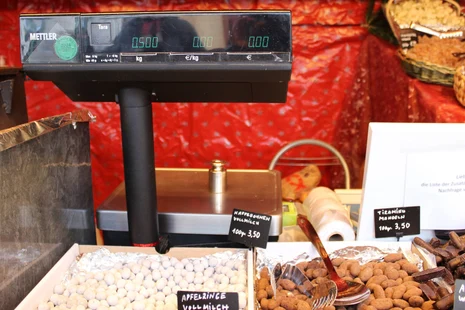 Waage an einem Wochen- oder Weihnachtsmarkt-Stand Auf einer Waage an einem Stand für schokolierte und karamellisierte Mandeln, Früchte und Kaffeebohnen steht ein 500-Gramm-Prüfgewicht. Die Anzeige zeigt 0,500 kg. Neben der Anzeige klebt ein grünes „geeicht bis 2024“-Kennzeichen.
