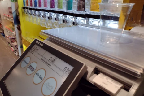 Kunden-Wage im Supermarkt zum selbstständigen Verwiegen von Süßigkeiten Foto einer Kunden-Wage im Supermarkt. Auf der Waage steht ein leerer Becher. Das Display zeigt 0 Gramm. Darunter sind vier Klickflächen zum Antippen, welche Ware man wiegen möchte. Im Hintergrund sieht verschiedene Süßigkeiten zum losen Verkauf.