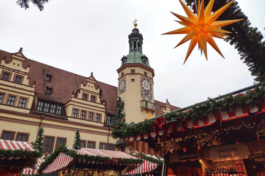 Blick vom Weihnachtsmarkt Leipzig zum Rathaus. Im Vordergrund sieht man festlich geschmückte Buden und einen Weihnachtsstern. Der vorderste Stand verkauft Südtiroler Feinkost und Wein.