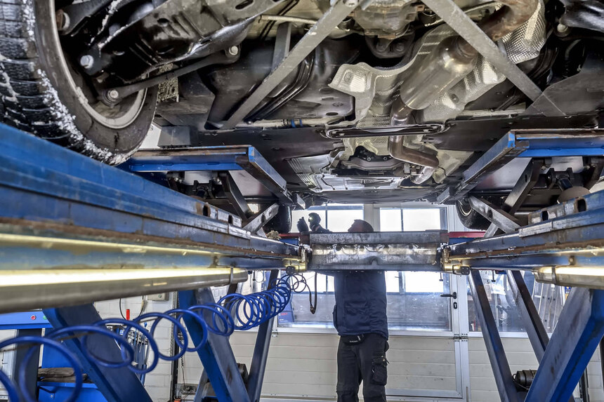 TÜV-Kontrolle: IM Hintergrund leichtet ein Mann mit einer Taschenlampe an das linke Vorderrad eines Fahrzeugs. Dieses steht auf einer Hebebühne, man sieht den Unterboden des Fahrzeugs.